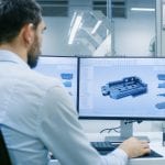 Engineer/ Technician Working on a Personal Computer with Two Displays, He's Designing New Component in CAD Program. Out of the Office Window Components Manufacturing Factory is Seen.