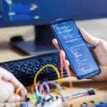 Developer is connecting breadboard to microcontroller. Man is holding smartphone with program code software for controlling electronic device. Chips, resistors, diodes on desktop of hardware engineer.