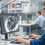 Over the Shoulder Shot of Engineer Working with CAD Software on Desktop Computer, Screen Shows Technical Details and Drawings. In the Background Engineering Facility Specialising on Industrial Design