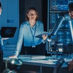 Diverse Team of Engineers with Laptop and a Tablet Analyse and Discuss How a Futuristic Robotic Arm Works and Moves a Metal Object. They are in a High Tech Research Laboratory with Modern Equipment.